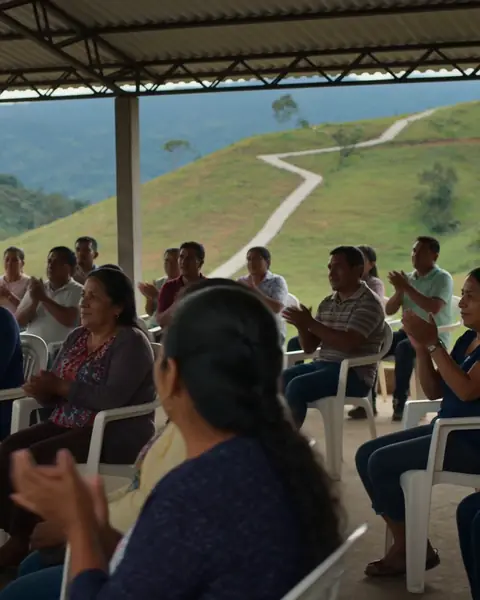 Reunión comunitaria al aire libre en una vereda de montaña; vecinos de distintas edades aplauden un camino recién hecho.
